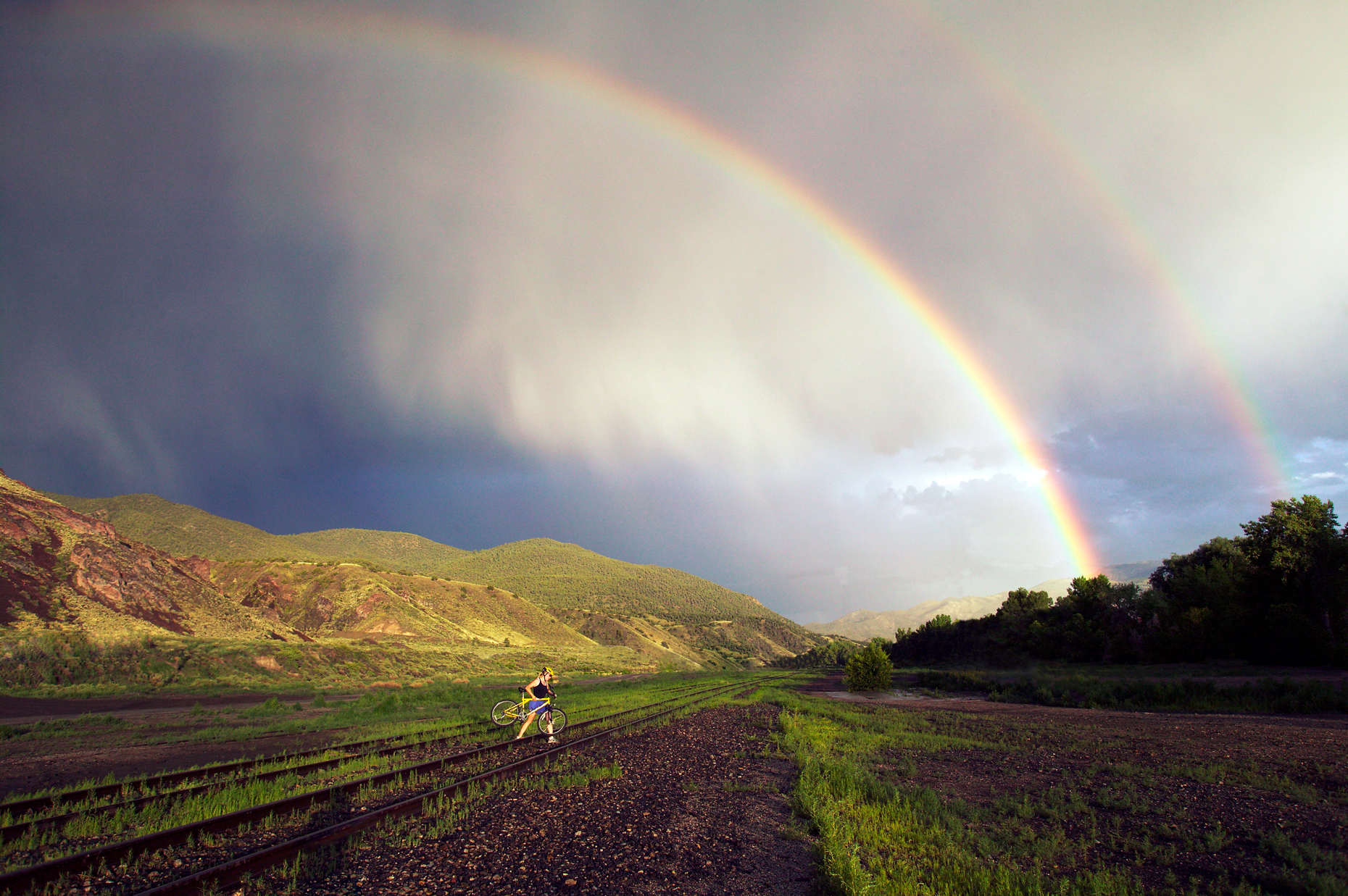 Rainbow over female mountain biker carrying her bike over railroad tracks to avoid a pending thunderstorm.